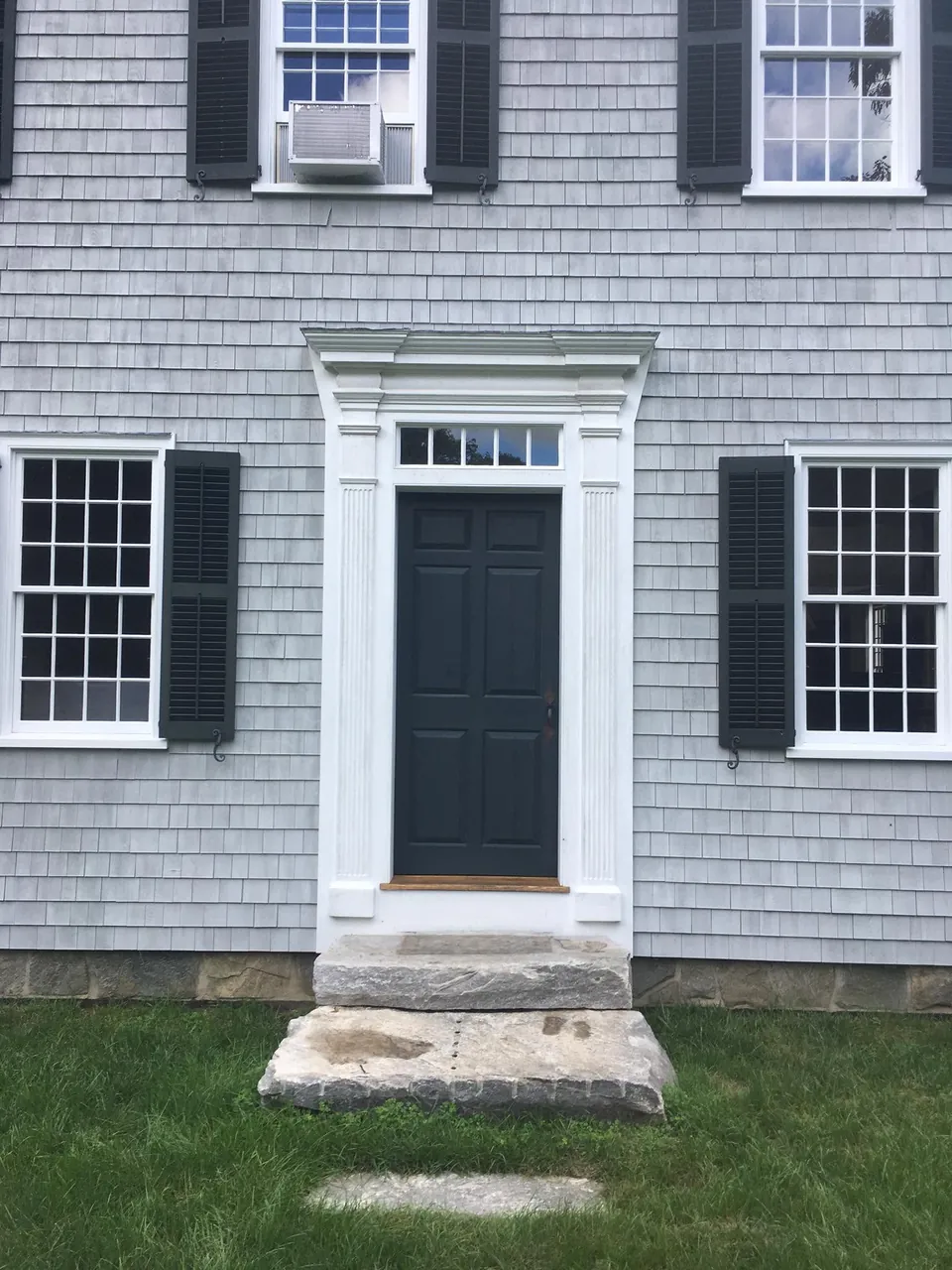 Dark green six-panel colonial entry door framed by fluted pilasters, dentil pediment, and a granite step
