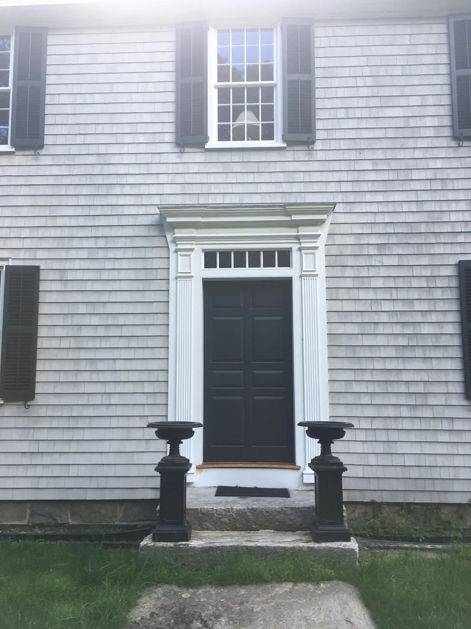 Black six-panel entry door with a pedimented surround and transom on a weathered cedar-shingled colonial